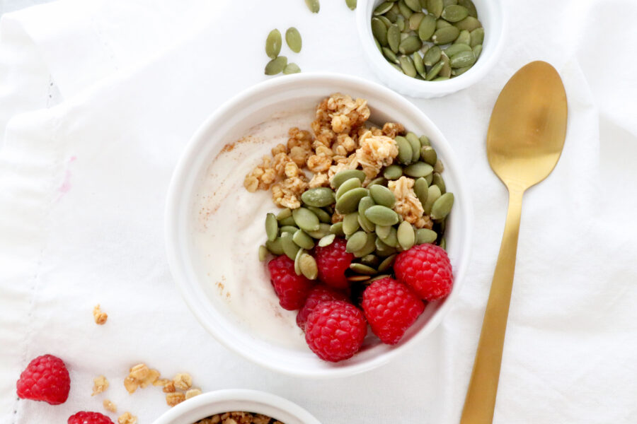 Bowl of cinnamon yogurt in center of photo topped with berries, granola and seeds. Bowl of pumpkin seeds above and fruit below. Gold spoon on the right side.