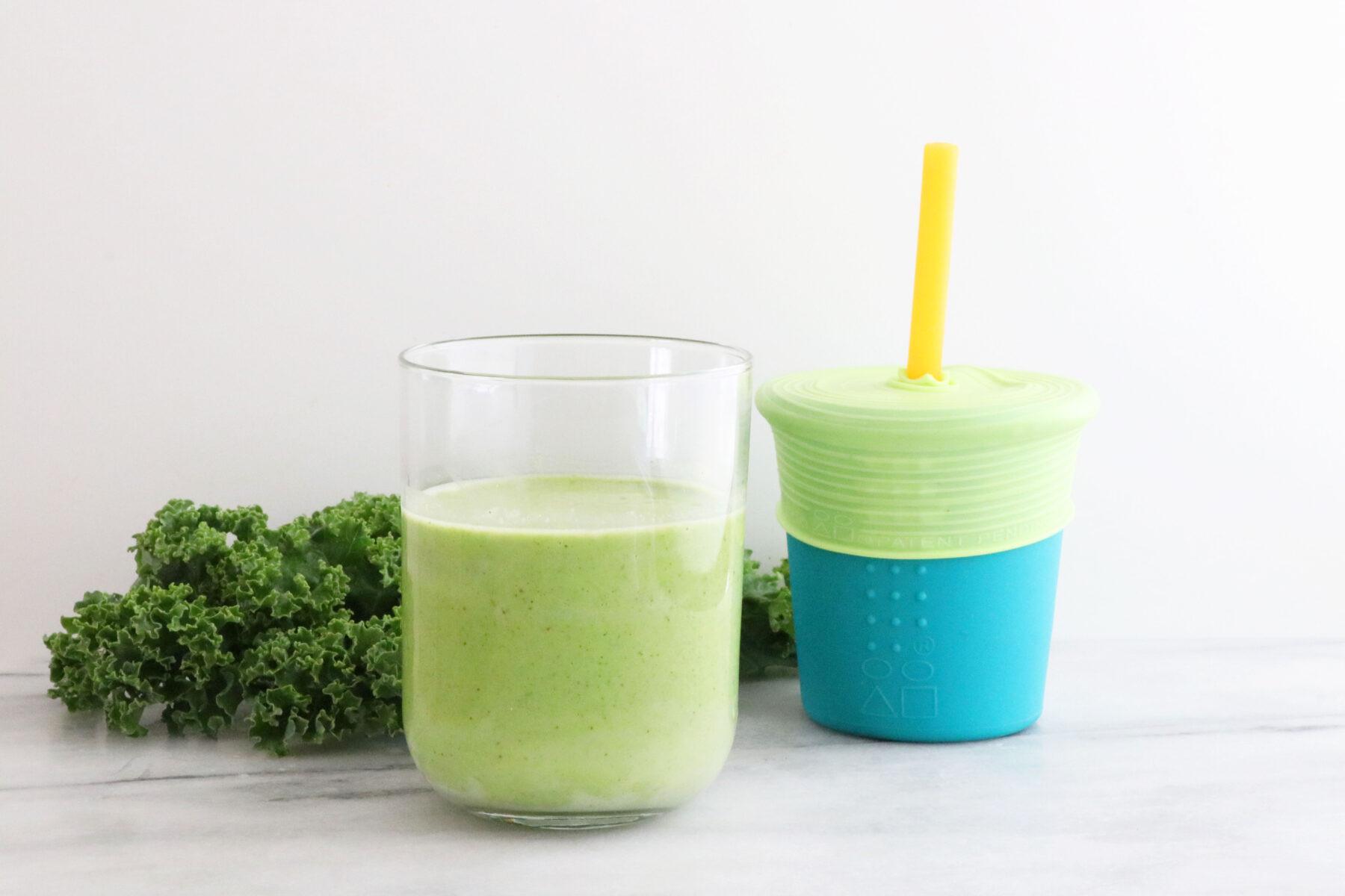 Kale on the left side, glass in the middle with smoothie and toddler cup on the right with smoothie and straw.
