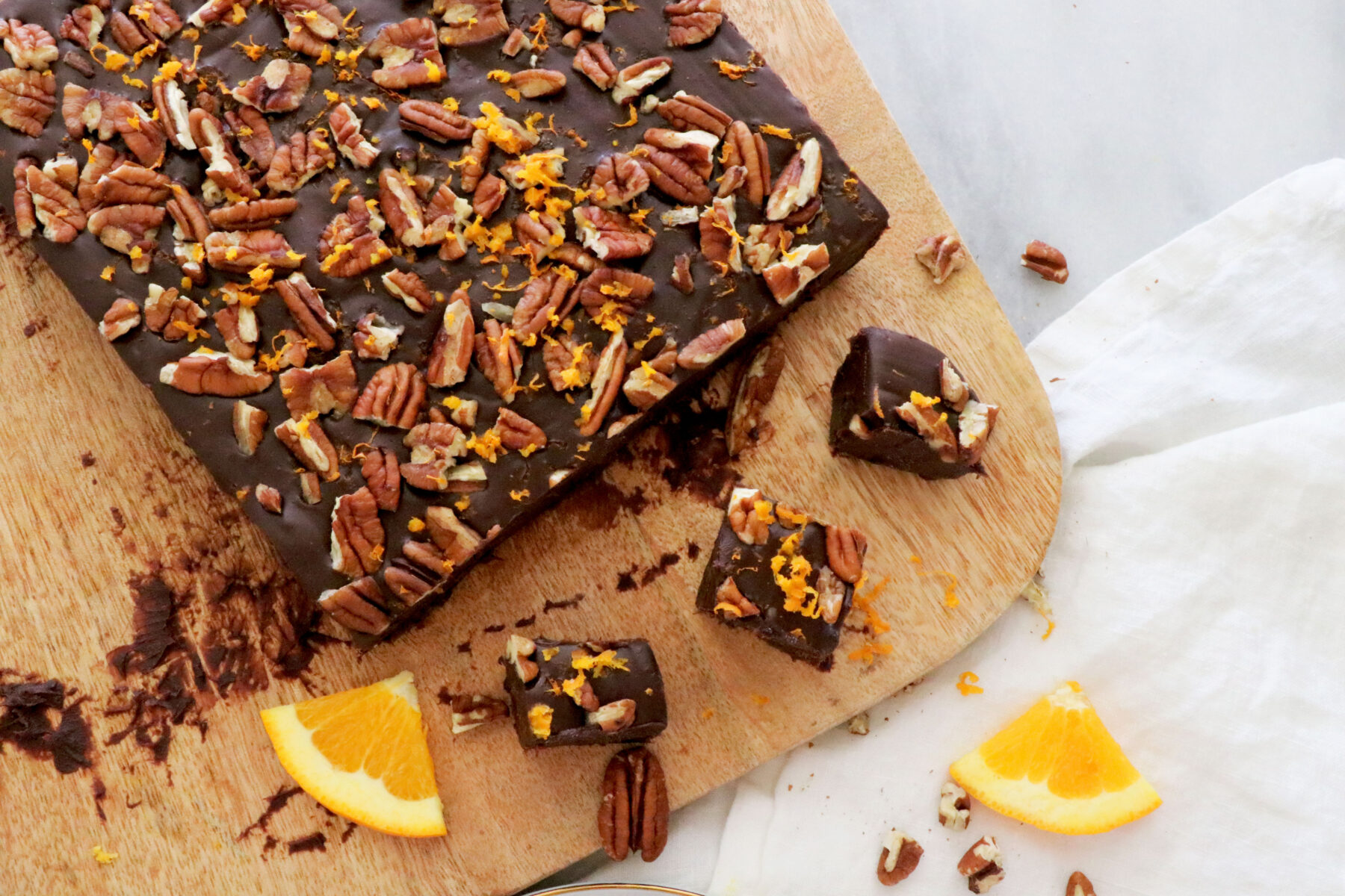 Wooden cutting board with chocolate orange fudge on top with a few squares cut out. Topped with pecans and orange zest