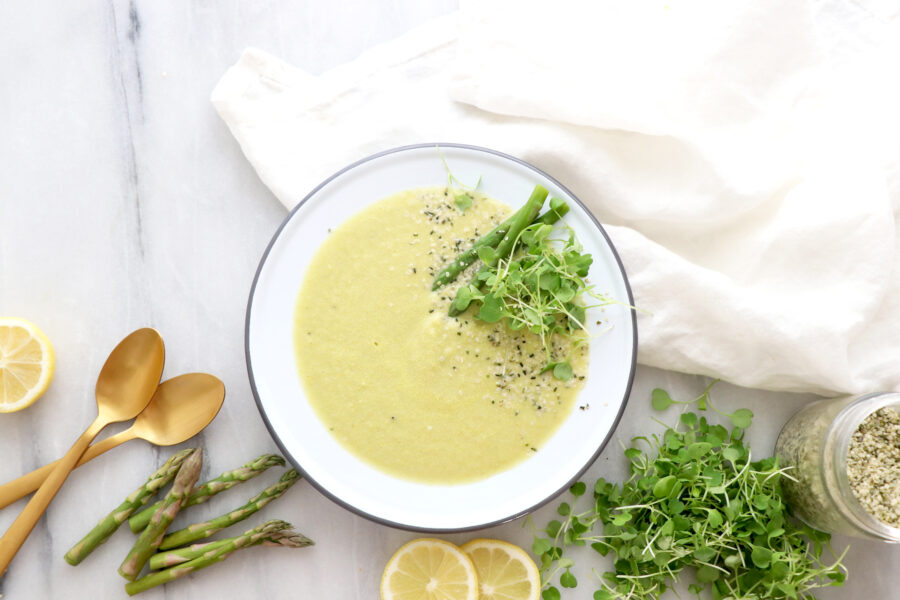Bowl of lemon asparagus soup with gold spoons, white napkin, and produce surrounding bowl.