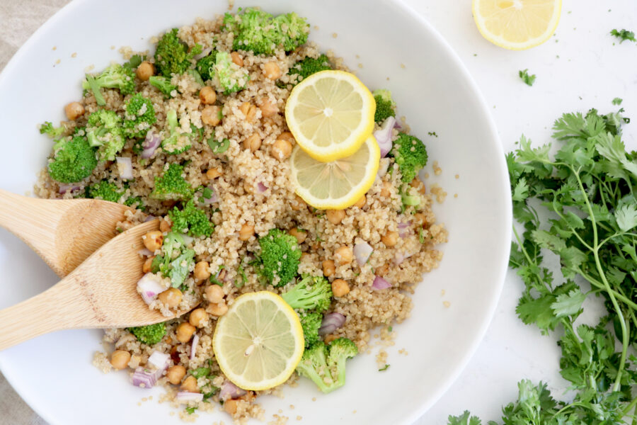 White bowl filled with quinoa, chickpea and broccoli salad. Salad tongs inside bowl and cilantro on the right side. 